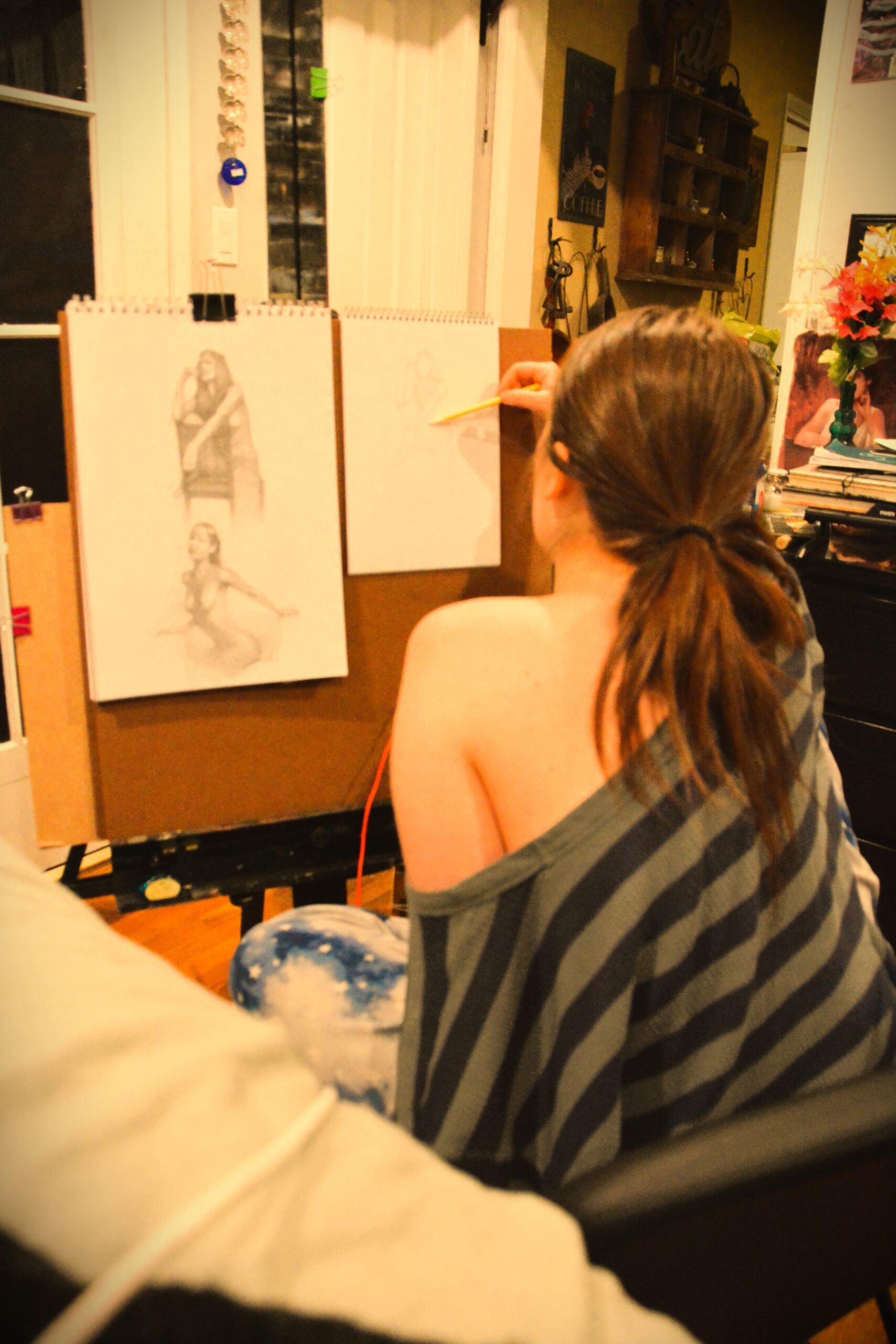 A photograph image of the backside of Samantha sitting on her favorite chair sketching a copy from her teacher's lesson of a woman sitting on a chair and a woman posing.